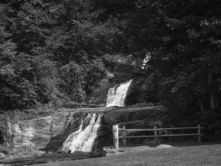Black-and-white photograph of Kent Falls cascading through woodland beside a wooden railing.