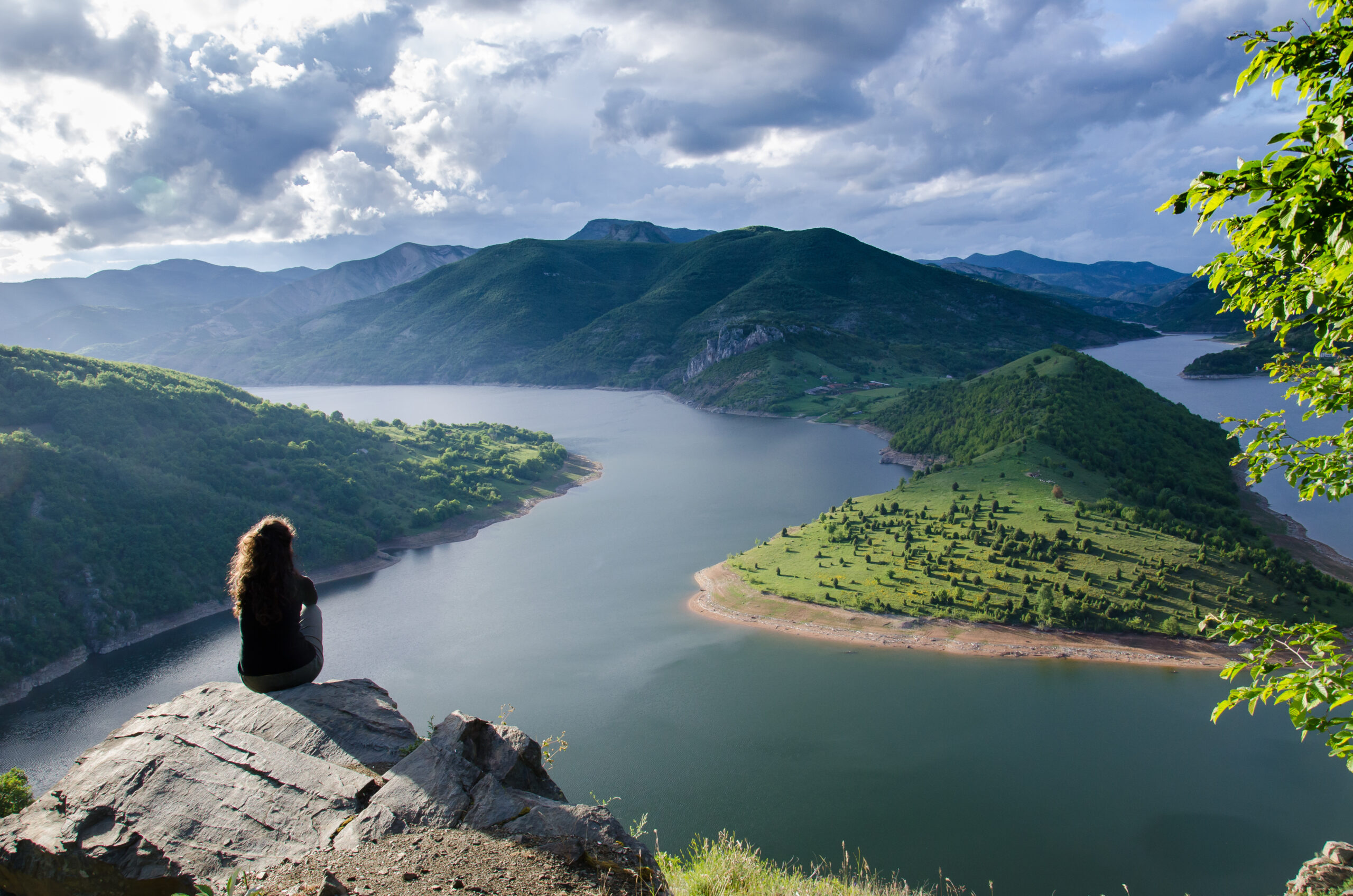 A person sitting on a rocky overlook above a lake surrounded by green mountains.