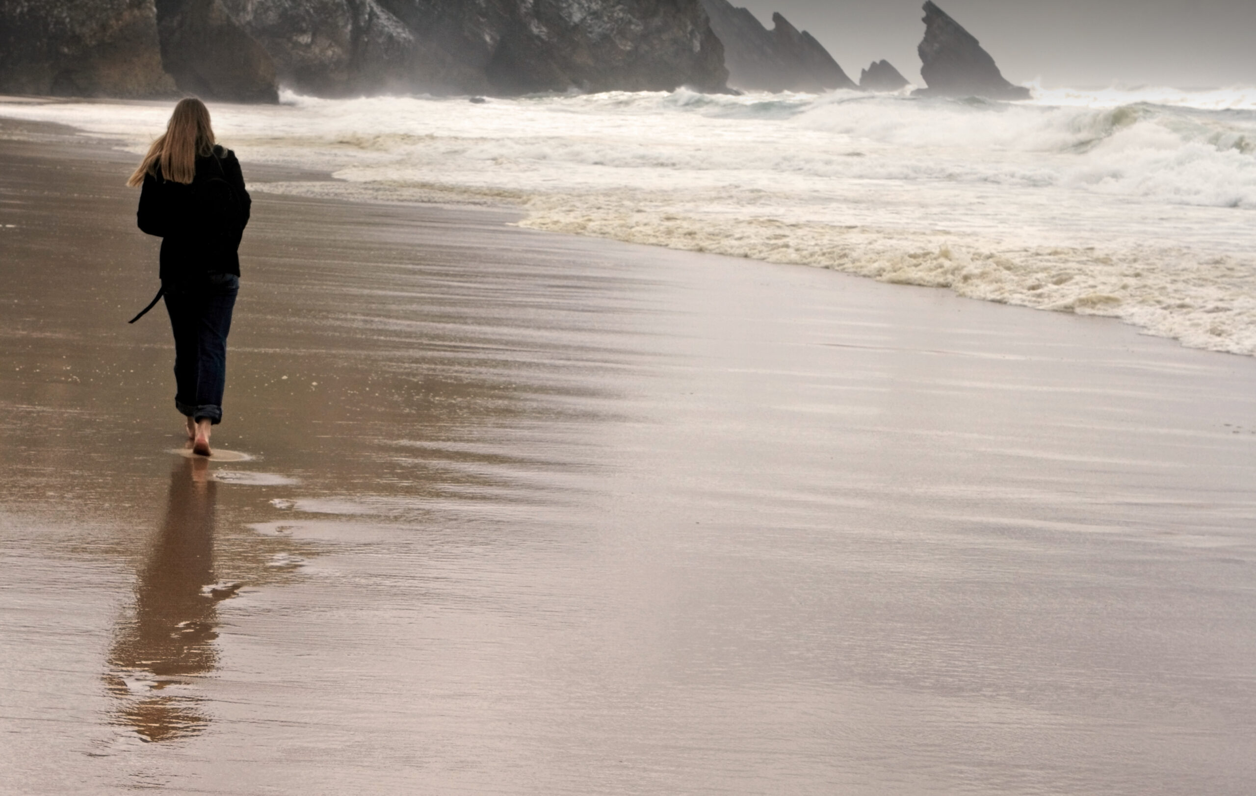 A woman walking along the shoreline beside breaking waves.