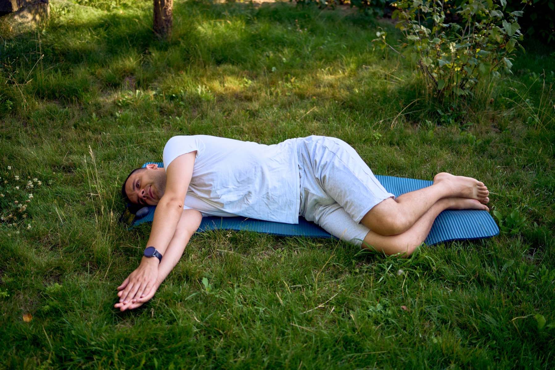 A man lying on his side on a mat in a garden, with bent knees and arms reaching forward.