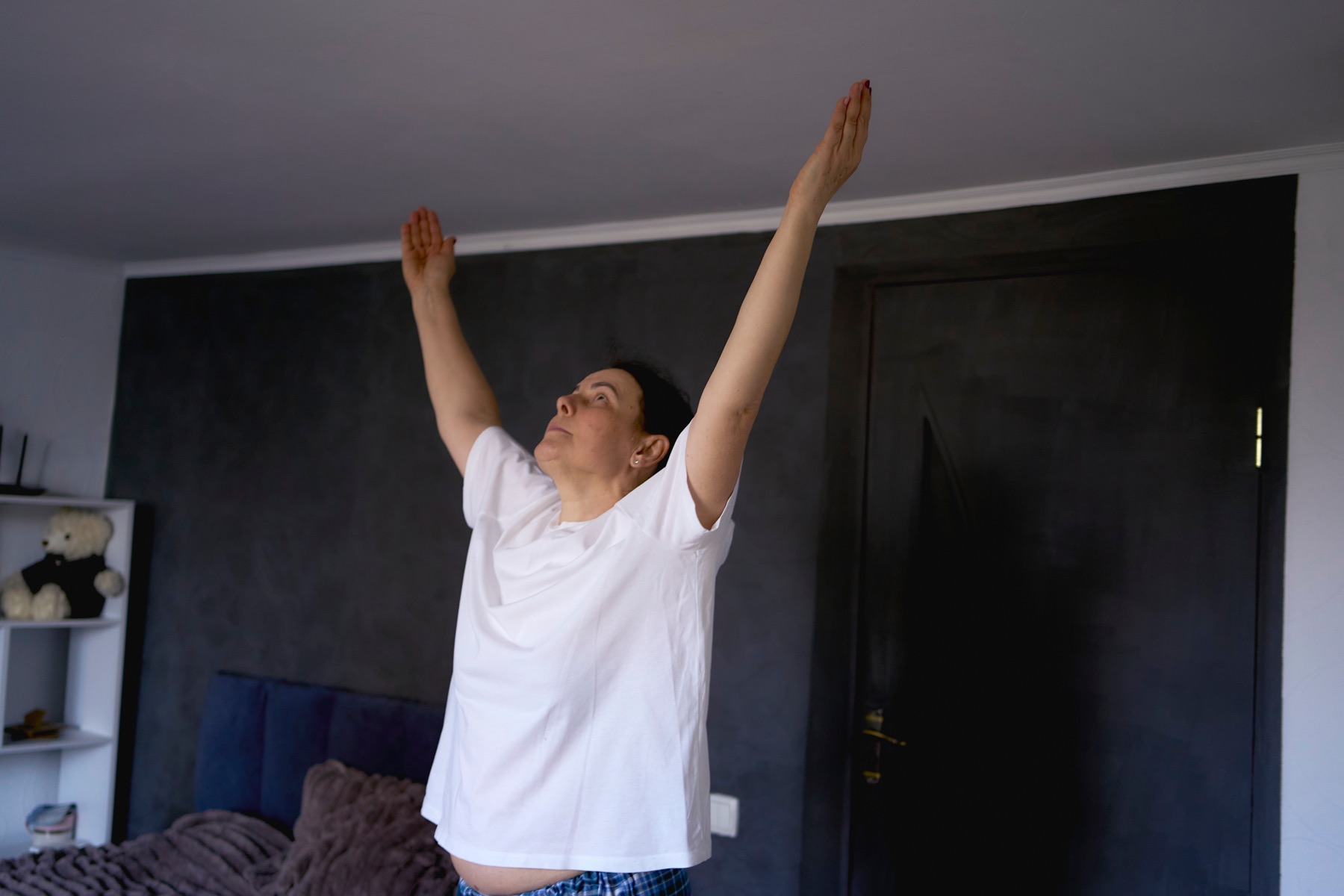 An older woman reaching both arms overhead while standing at home.