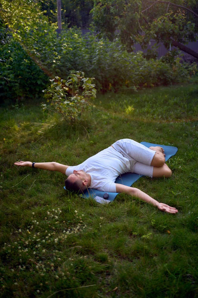 Man lying on a mat in a garden with arms open and bent knees resting to one side.
